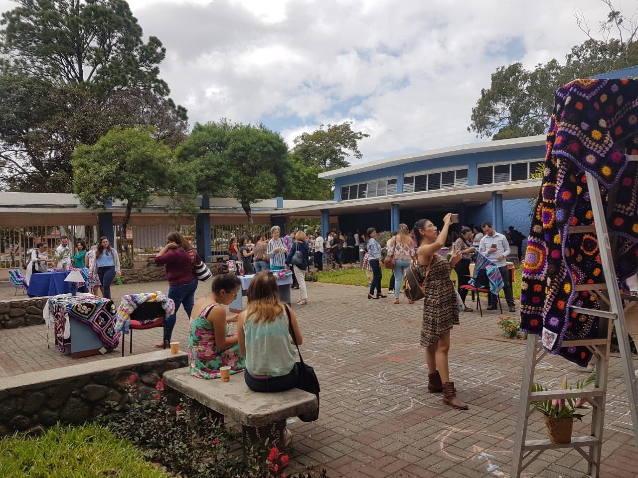 Los participantes de la jornada se encuentran en el patio central de la Facultad de Educación, algunos sentados en bancas, otros de pie. Los rodean los trabajos de la artista Estrella Fages, éstos son piezas tejidas de diferentes colores y tamaños.