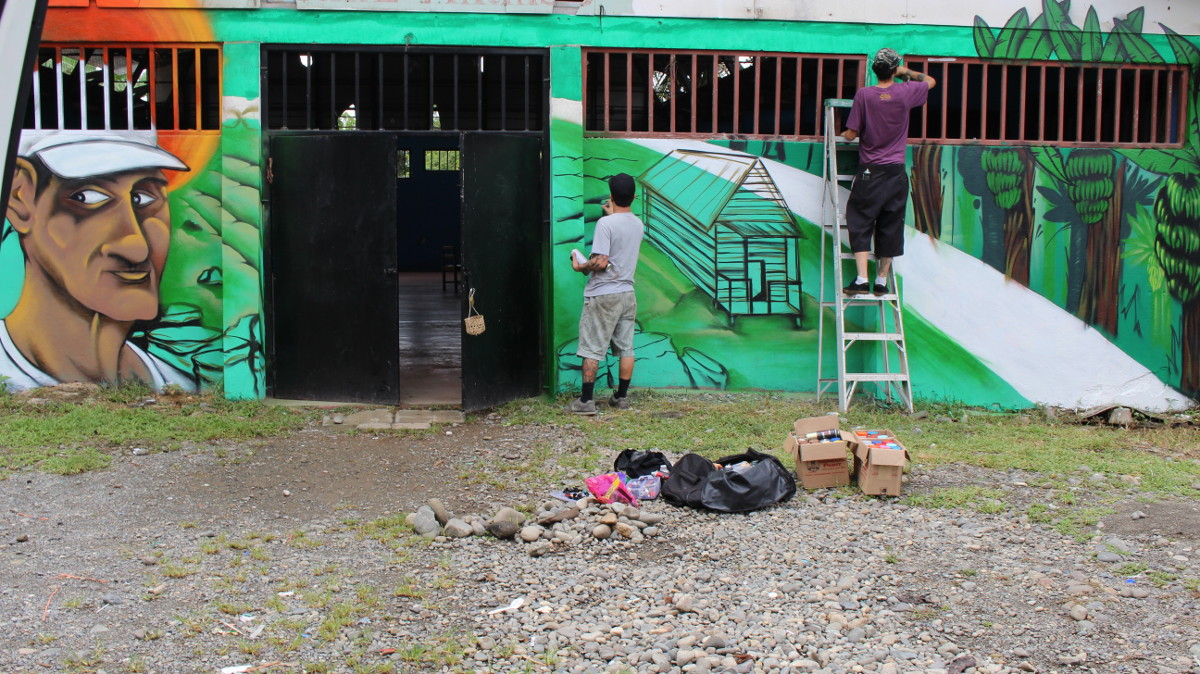 Los artistas Mush y Kein de espaldas trabajando sobre la pared. A la izquierda está la imagen de perfil del líder comunitario “don Roberto”. Fotografía del Programa Kioscos Socio-ambientales.