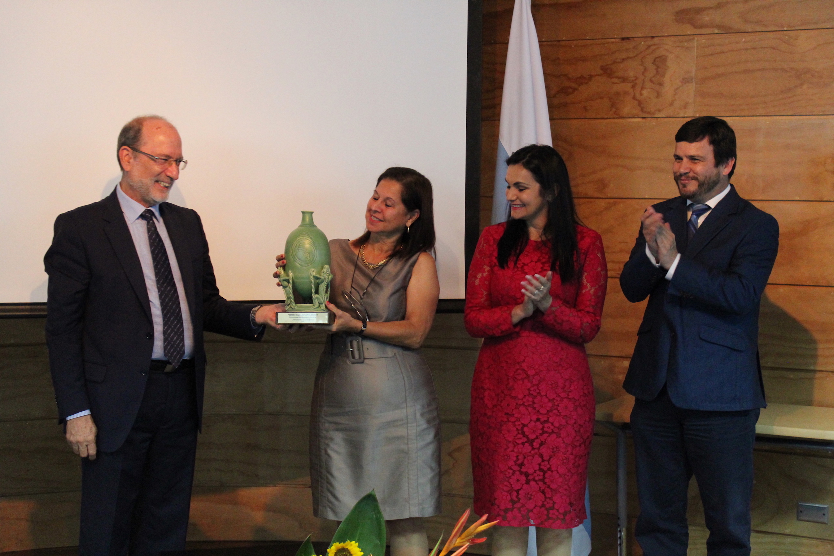 La Dra. Helga Blanco recibe la estatuilla color verde en forma vasija del Rector Henning Jensen. A su izquierda, se encuentran la vicerrectora de Acción Social, Marjorie Jiménez, y el presidente del Consejo Universitario, Rodrigo Carboni. Los cuatro se encuentran frente a la pantalla principal del Auditorio de Educación Continua. 